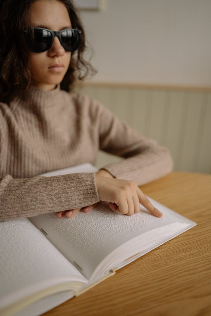 A visually impaired woman reading a Braille book at a desk indoors, focusing on accessibility and disability awareness.
